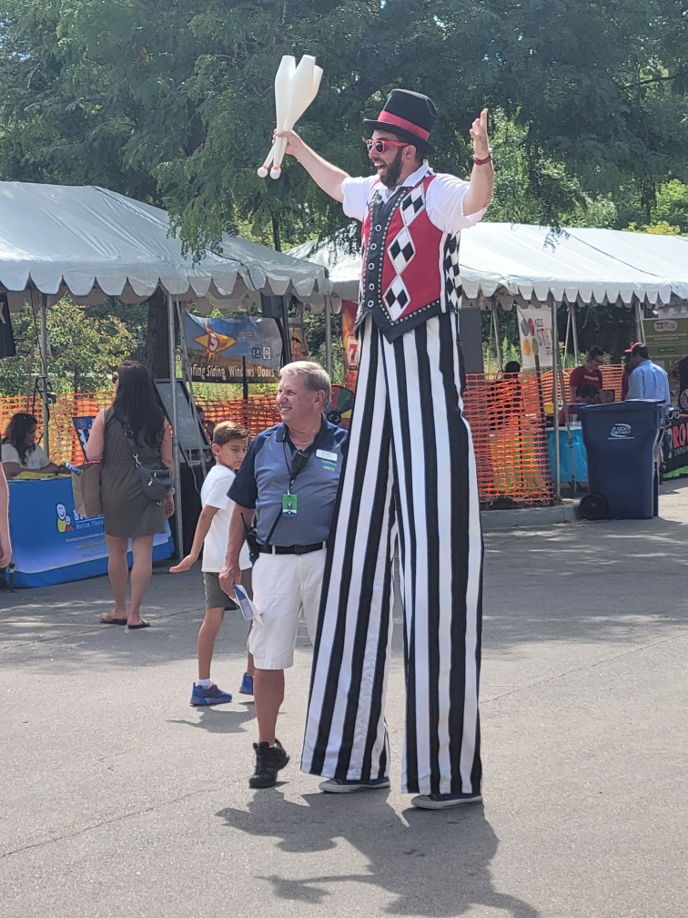 Photo by Joslyn Talken. A man standing on stilts smiling while holding bowling pins with several stands in the background